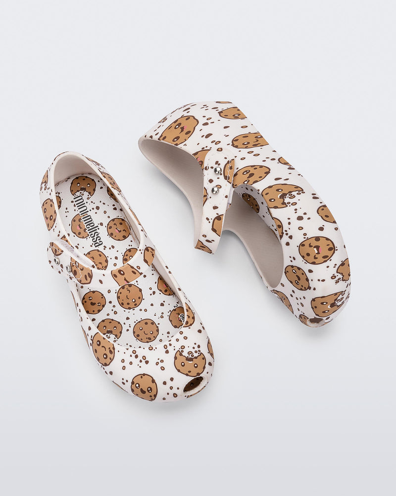 Top and side view of the Mini Melissa Ultragirl flats in candy print. The print is an all over white / beige chocolate chip cookie print. The shoe has a single velcro strap across the front of the foot. 