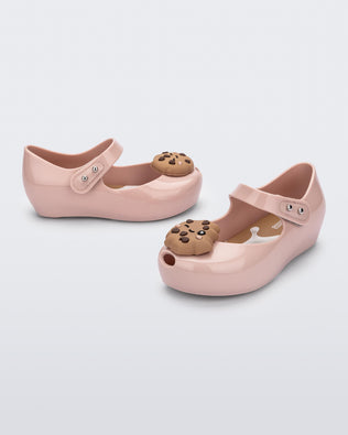 Angled view of a pair of Mini Melissa Ultragirl Candy ballet flats in Pink colorway. The shoe has a single strap across the top of the foot with a double snap closure. The product also has a 3D chocolate chip cookie applique on the top fo the toe. The cookie applicque is a light brown color with darker brown colored chips. 