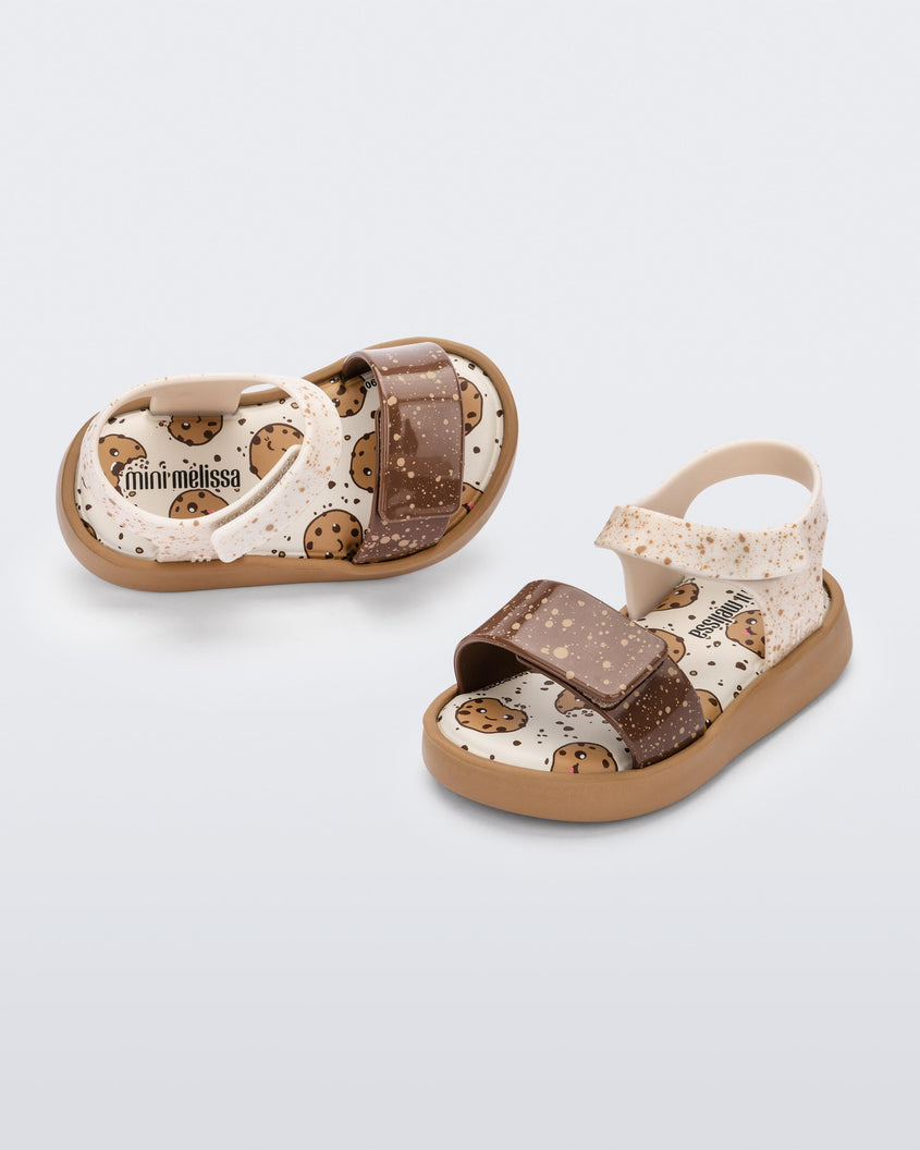 An angled top and side view of a pair of beige/brown Mini Melissa Jump Candy sandal with white and brown crumb detail straps and a print of cookies on the insole.