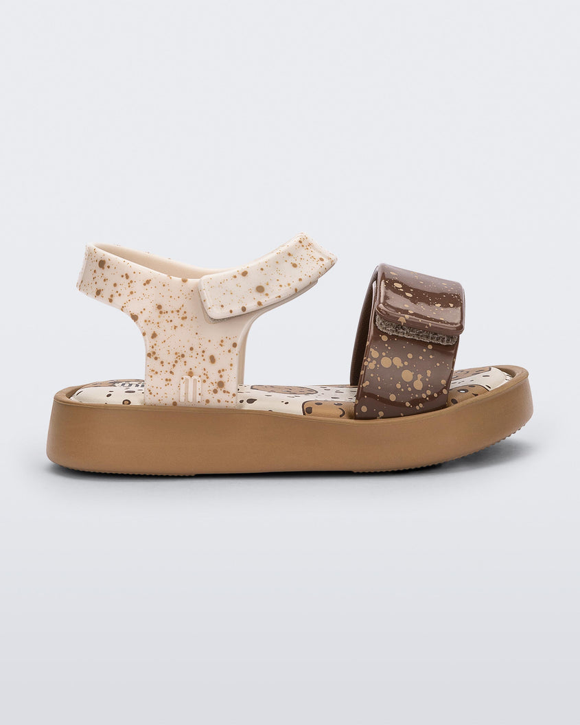 Side view of a beige/brown Mini Melissa Jump Candy sandal with white and brown crumb detail straps and a print of cookies on the insole.
