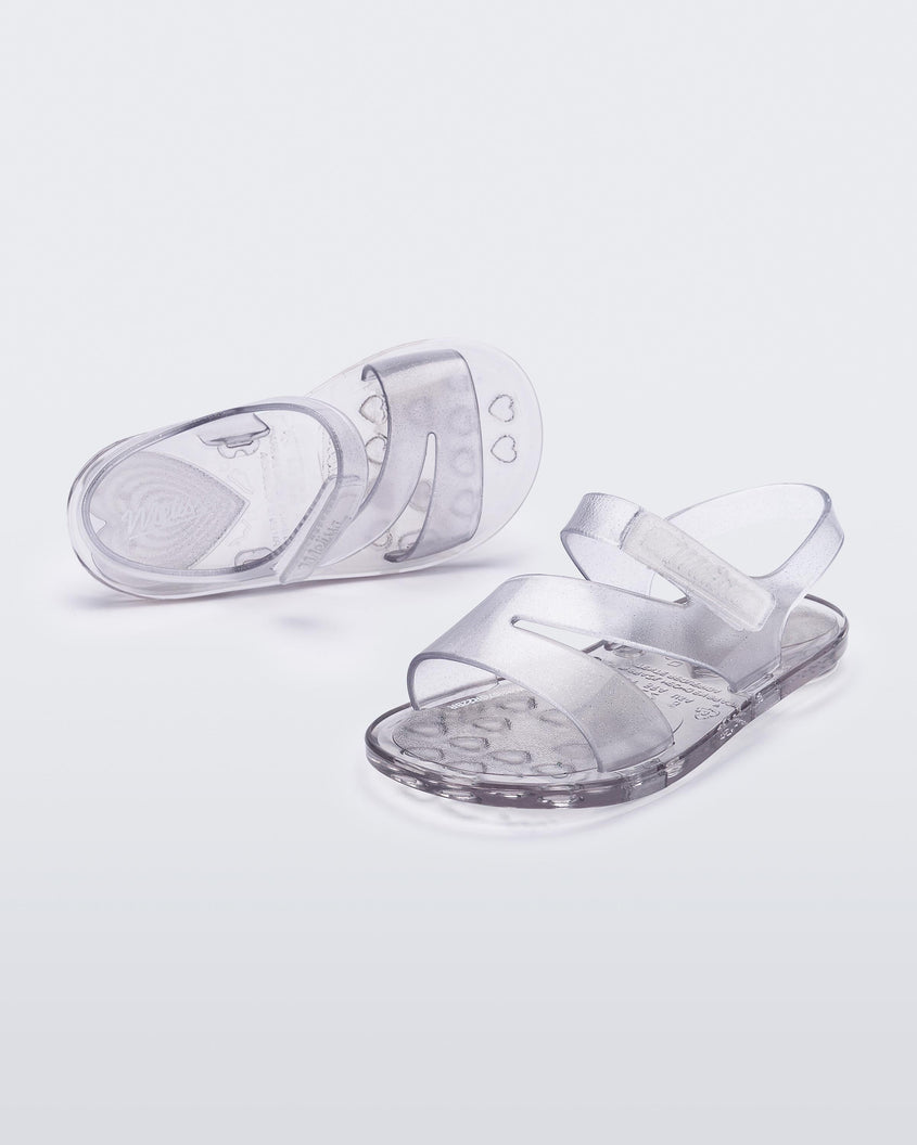 An angled front and top view of a pair of clear Mini Melissa The Real Jelly Paris sandals with three front straps and an ankle strap.