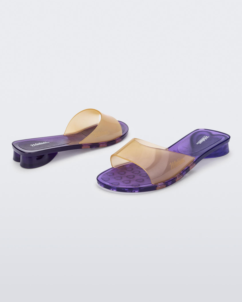 An angled front and side view of a pair of transparent purple/yellow Melissa Kim slides.