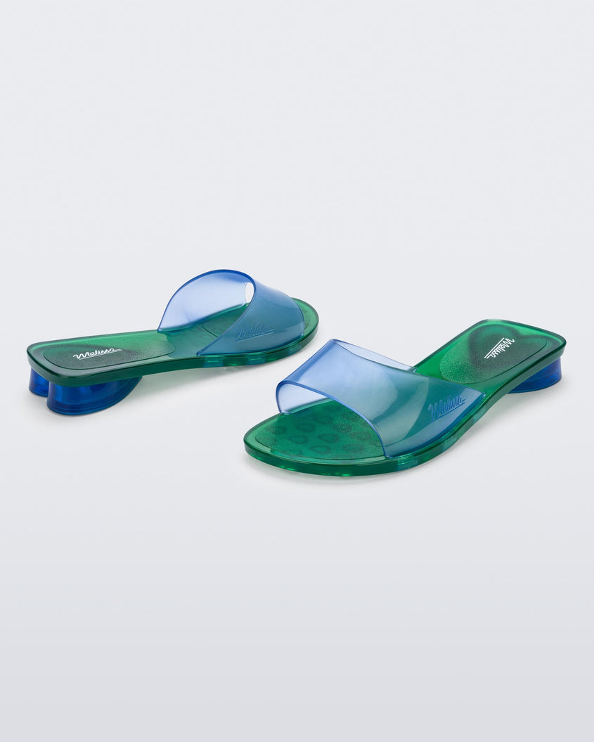 An angled front and side view of a pair of transparent blue/green Melissa Kim slides.