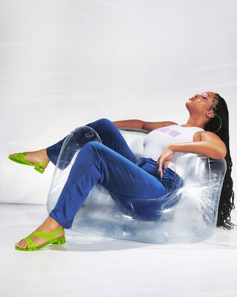 A model sitting in a clear chair, posing for a picture in a white top, jeans and a pair of green Melissa The Real Jelly Paris sandals with two front straps and an ankle strap.