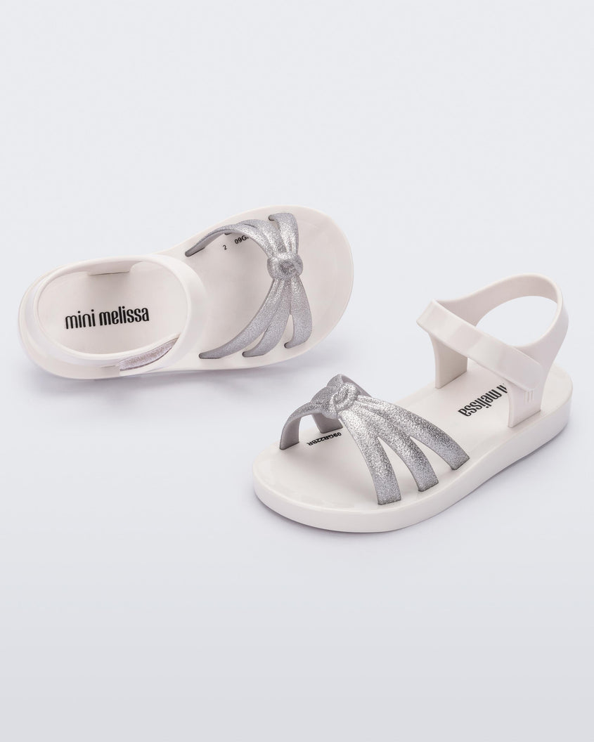 An angled front and top view of a pair of white/silver Mini Melissa Precious sandals with three silver glitter front straps joining in the middle and a white ankle strap.