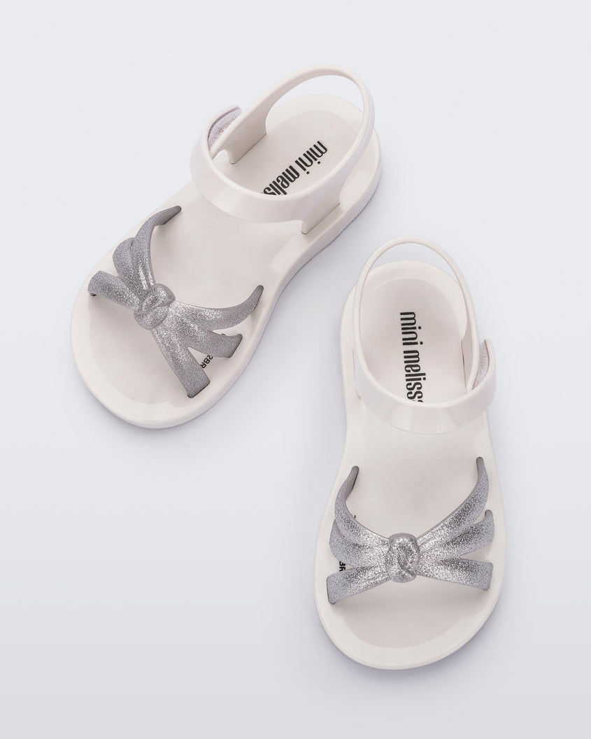 Top view of a pair of white/silver Mini Melissa Precious sandals with three silver glitter front straps joining in the middle and a white ankle strap.