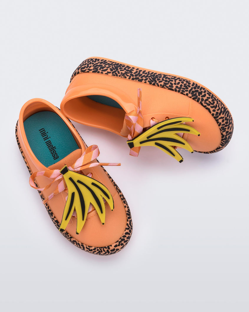 A top and side view of a pair of orange Mini Melissa Street sneakers with an orange base, black and orange patterned sole, orange and pink patterned laces with a drawing of bananas on top the laces.