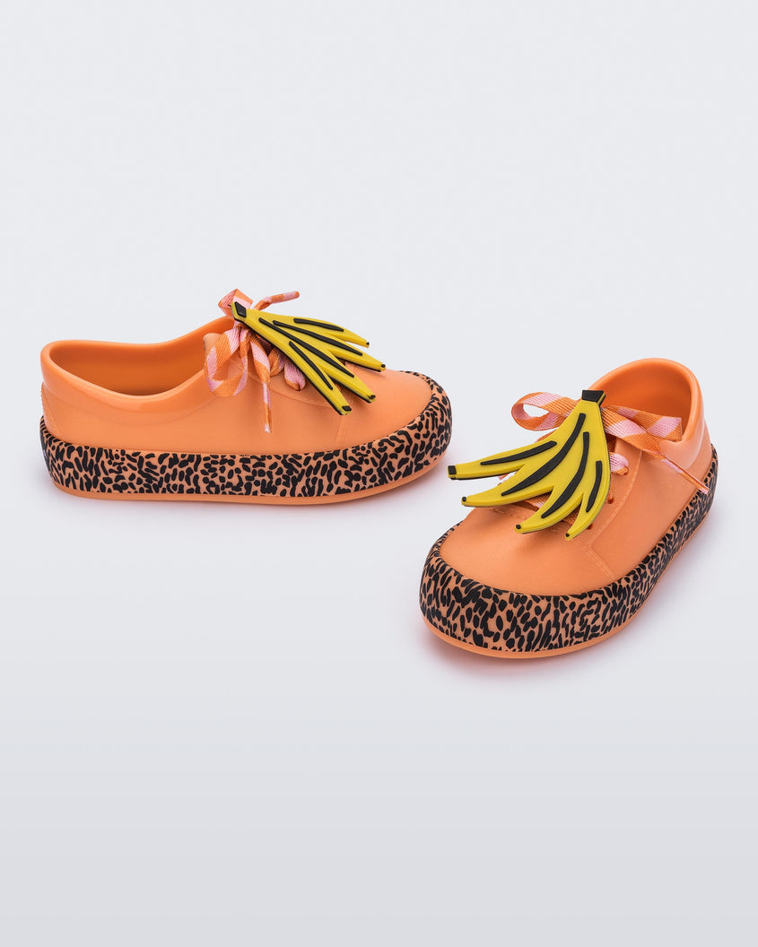 An angled front and side view of a pair of orange Mini Melissa Street sneakers with an orange base, black and orange patterned sole, orange and pink patterned laces with a drawing of bananas on top the laces.
