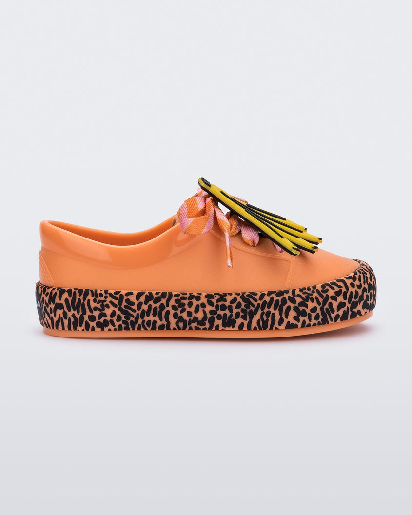Side view of a orange Mini Melissa Street sneaker with an orange base, black and orange patterned sole, orange and pink patterned laces with a drawing of bananas on top the laces.
