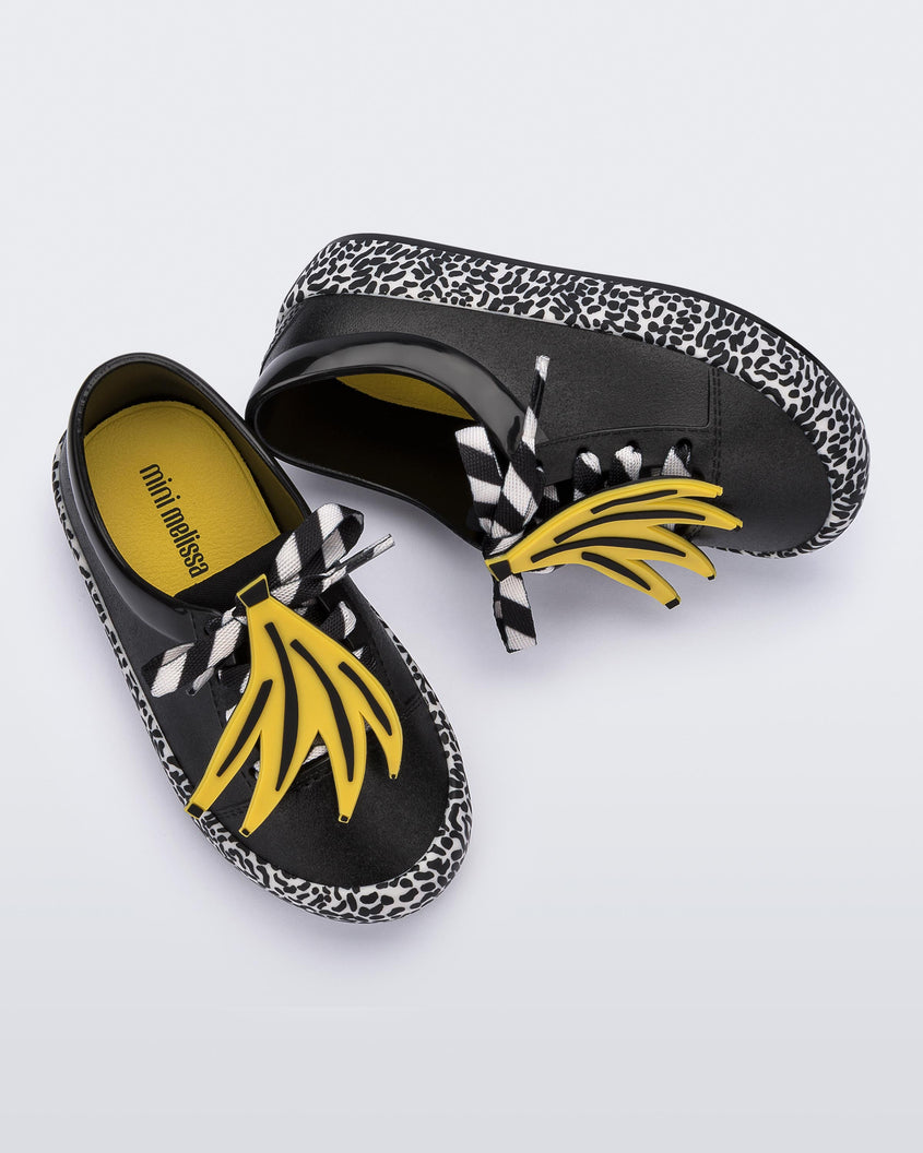 A top and side view of a pair of black Mini Melissa Street sneakers with an black base, black and white patterned sole, white and black patterned laces with a drawing of bananas on top the laces.