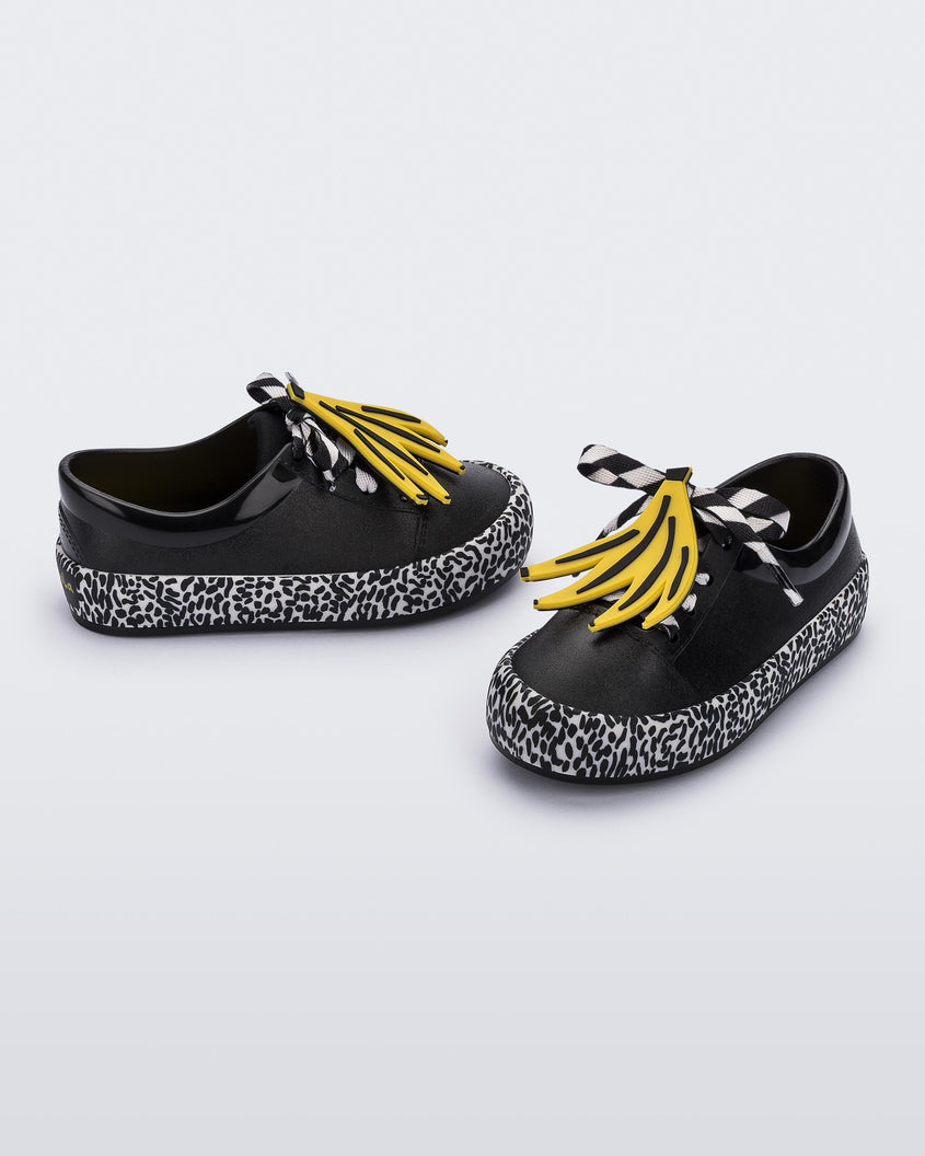 An angled front and side view of a pair of black Mini Melissa Street sneakers with an black base, black and white patterned sole, white and black patterned laces with a drawing of bananas on top the laces.
