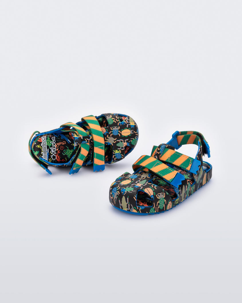 An angled front and top view of a pair of blue/green Mini Melissa Yoyo sandals with a black, green and blue patterned base, with two green and orange velcro front straps and an ankle strap.