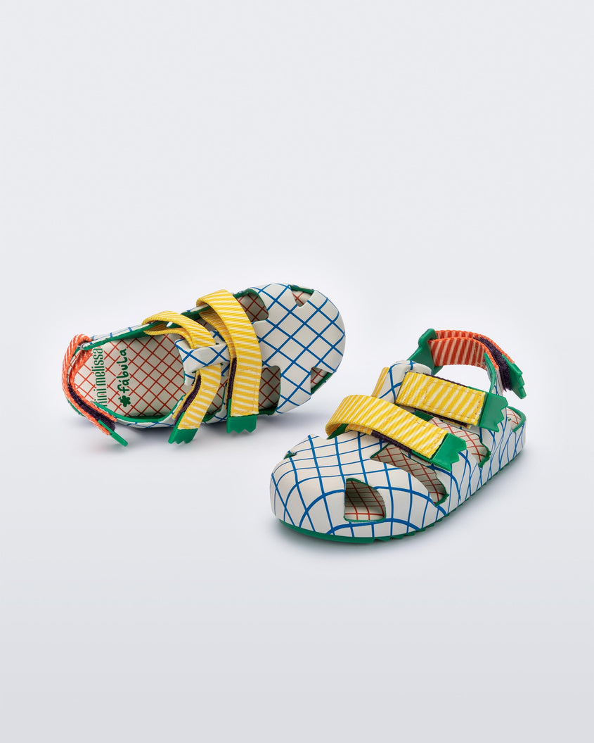 An angled front and top view of a pair of green/beige Mini Melissa Yoyo sandals with a gray, green and blue patterned base, with two green and yellow velcro front straps and a red and green ankle strap.