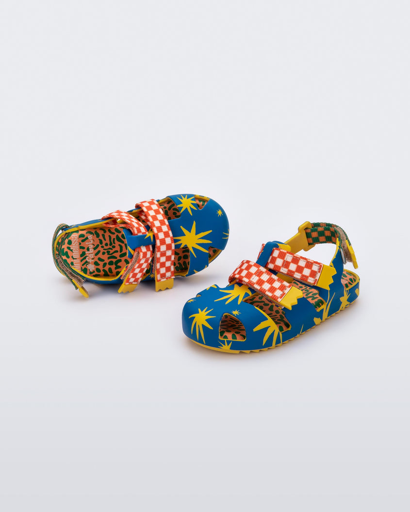 An angled side and top view of a pair of yellow/blue Mini Melissa Yoyo sandals with a yellow and blue patterned base, with two red and white velcro front straps and an orange and green ankle strap.