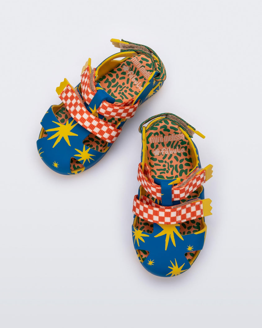 Top view of a pair of yellow/blue Mini Melissa Yoyo sandals with a yellow and blue patterned base, with two red and white velcro front straps and an orange and green ankle strap.