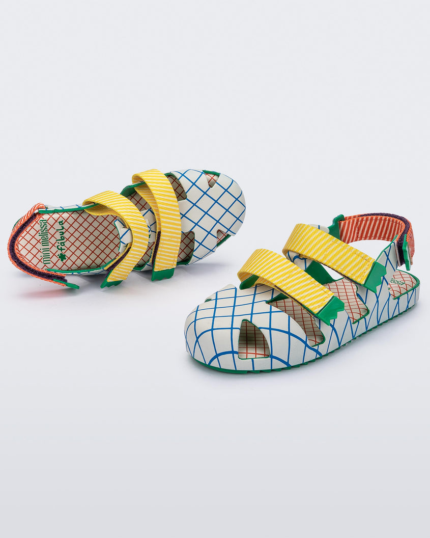 An angled side and top view of a pair of Green/Beige Mini Melissa Yoyo sandals with a gray/blue patterned base with two front yellow and green velcro straps and a red and green ankle strap.