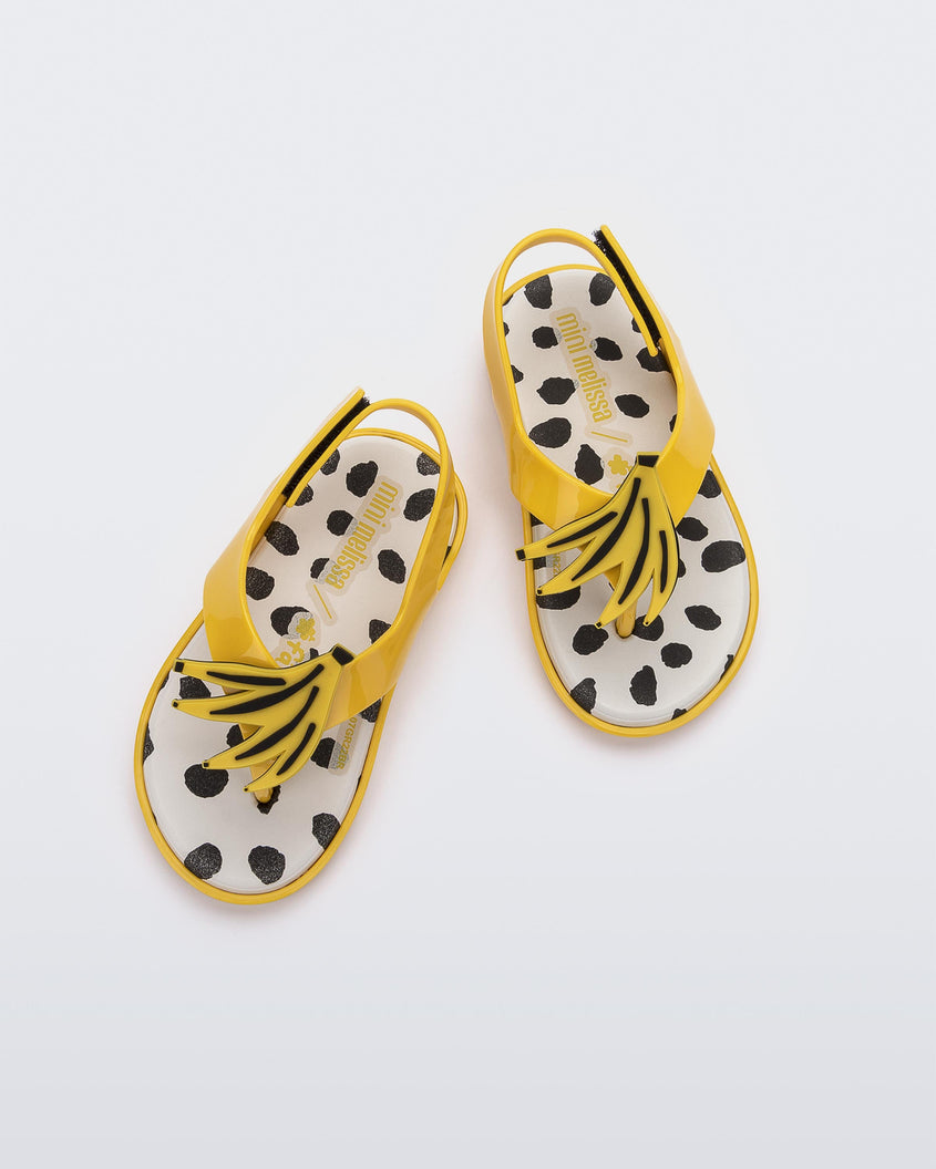 Top view of a pair of yellow Mini Melissa Sunny sandals with a black base, a banana drawing on top of the straps and a black and white polka dot insole.