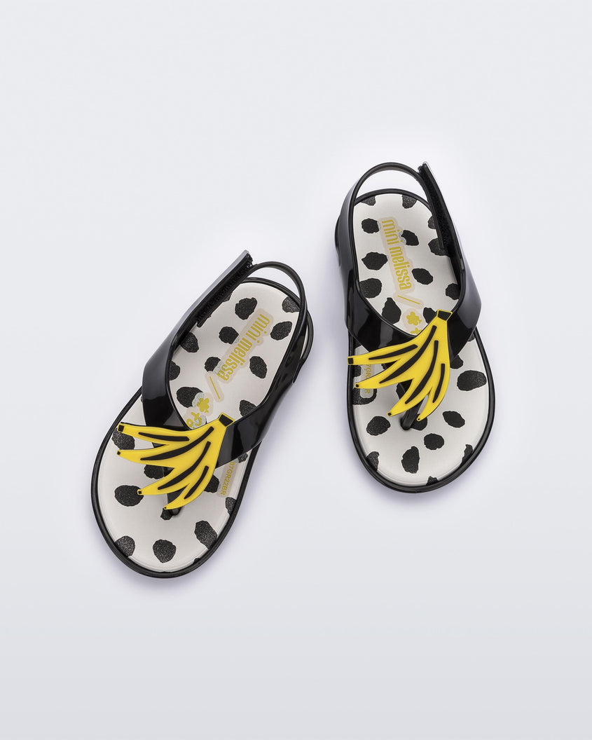 Top view of a pair of black Mini Melissa Sunny sandals with a black base, a banana drawing on top of the straps and a black and white polka dot insole.