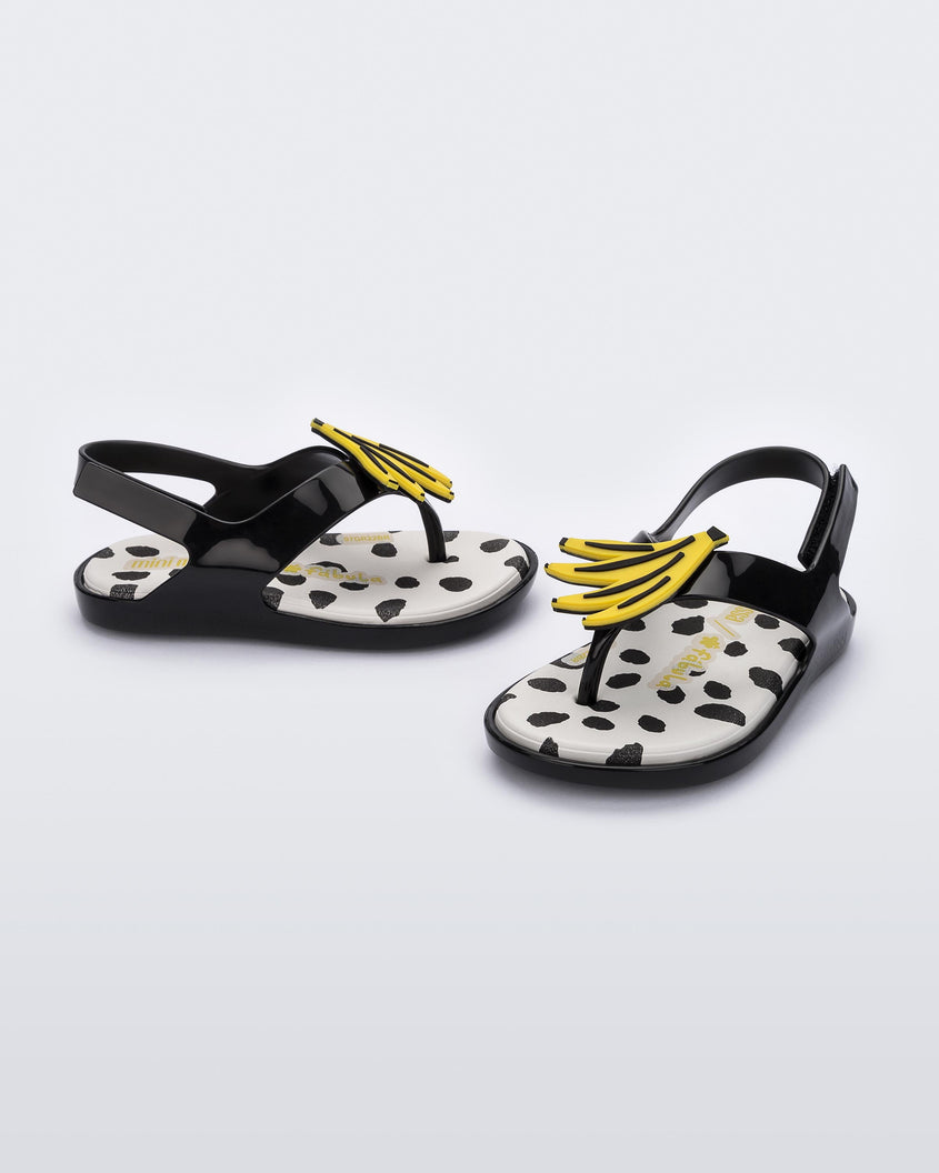 An angled front and side view of a pair of black Mini Melissa Sunny sandals with a black base, a banana drawing on top of the straps and a black and white polka dot insole.