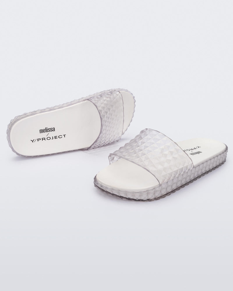 An angled top and side view of a pair of transparent clear/white Melissa Court Slides with a checkered pattern texture.