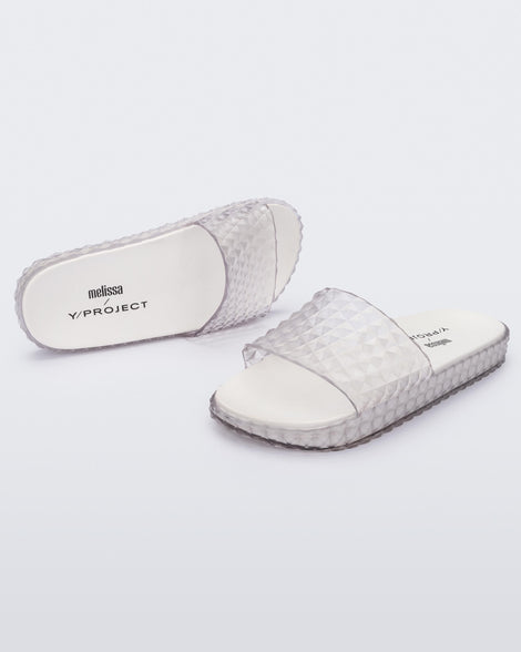 An angled top and side view of a pair of transparent clear/white Melissa Court Slides with a checkered pattern texture.
