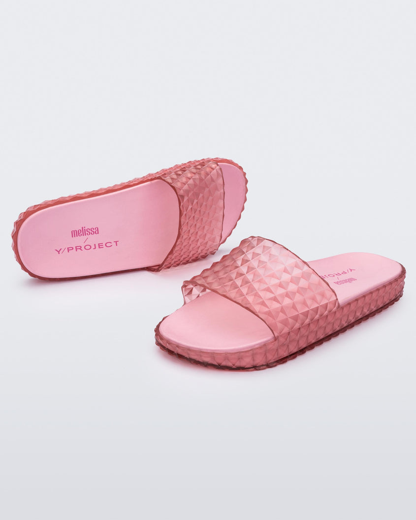 An angled front and side view of a pair of transparent pink Melissa Court Slides with a checkered pattern texture.