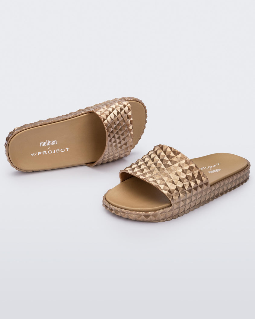 An angled top and side view of a pair of gold Melissa Court Slides with a checkered pattern texture.