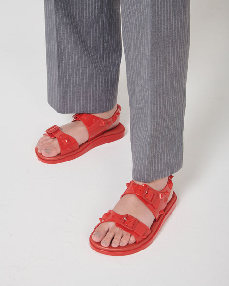A model's legs in gray pants and a pair of red Melissa Spikes Sandals with two straps with buckles on top and spike details around the base.