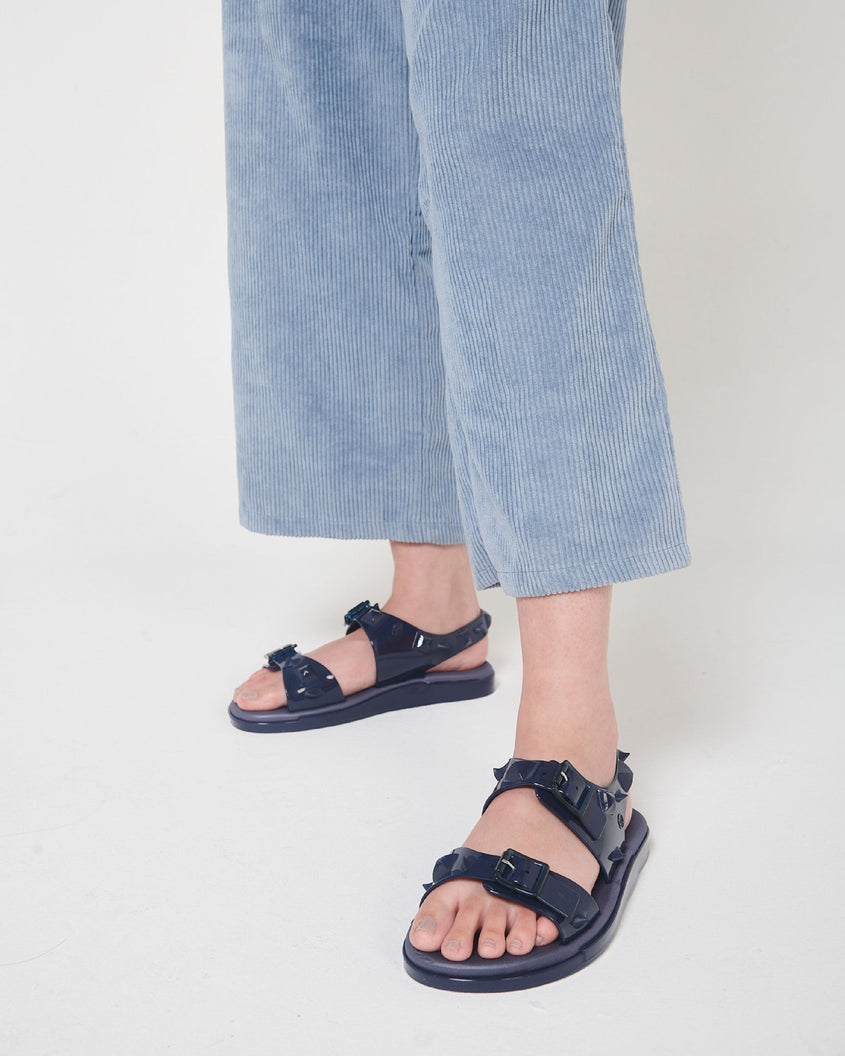 A model's legs in blue corduroy pants and a pair of blue Melissa Spikes Sandals with two straps with buckles on top and spike details around the base.