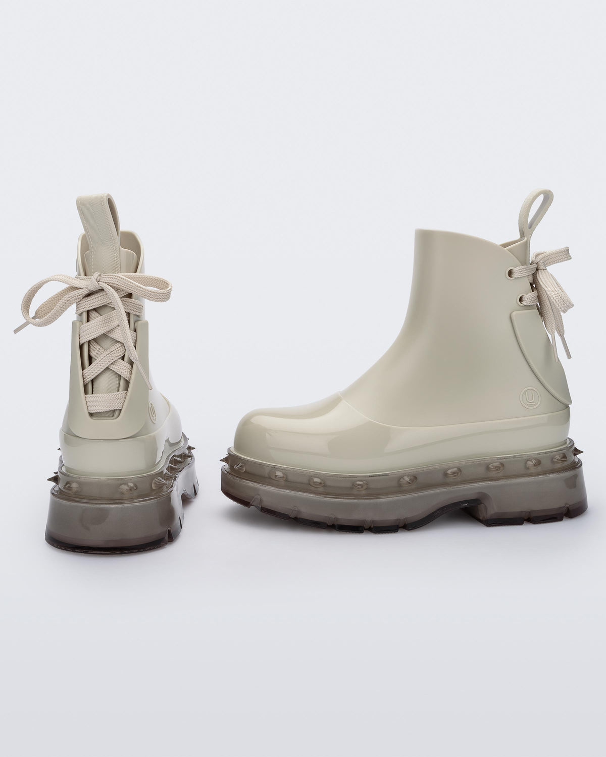 An angled side and back view of a pair of beige Melissa Spikes Boots with spike details around the sole and laces in the back.