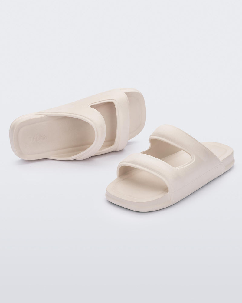 An angled front and side view of a pair of beige Melissa Free Grow slides with a square cut out on the front.