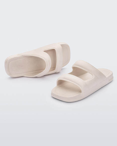 An angled front and side view of a pair of beige Melissa Free Grow slides with a square cut out on the front.