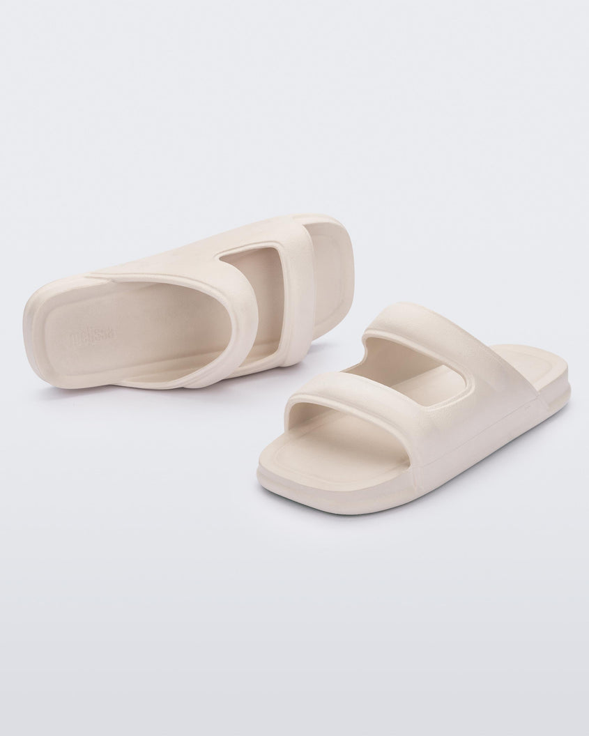 An angled front and side view of a pair of beige Melissa Free Grow slides with a square cut out on the front.
