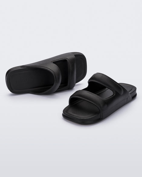 An angled front and side view of a pair of black Melissa Free Grow slides with a square cut out on the front.