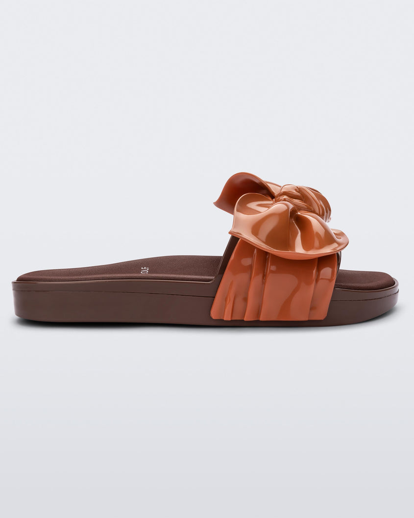 Side view of a Melissa Tie Beach slide with brown sole and brown/orange strap with 3D bow detail. 