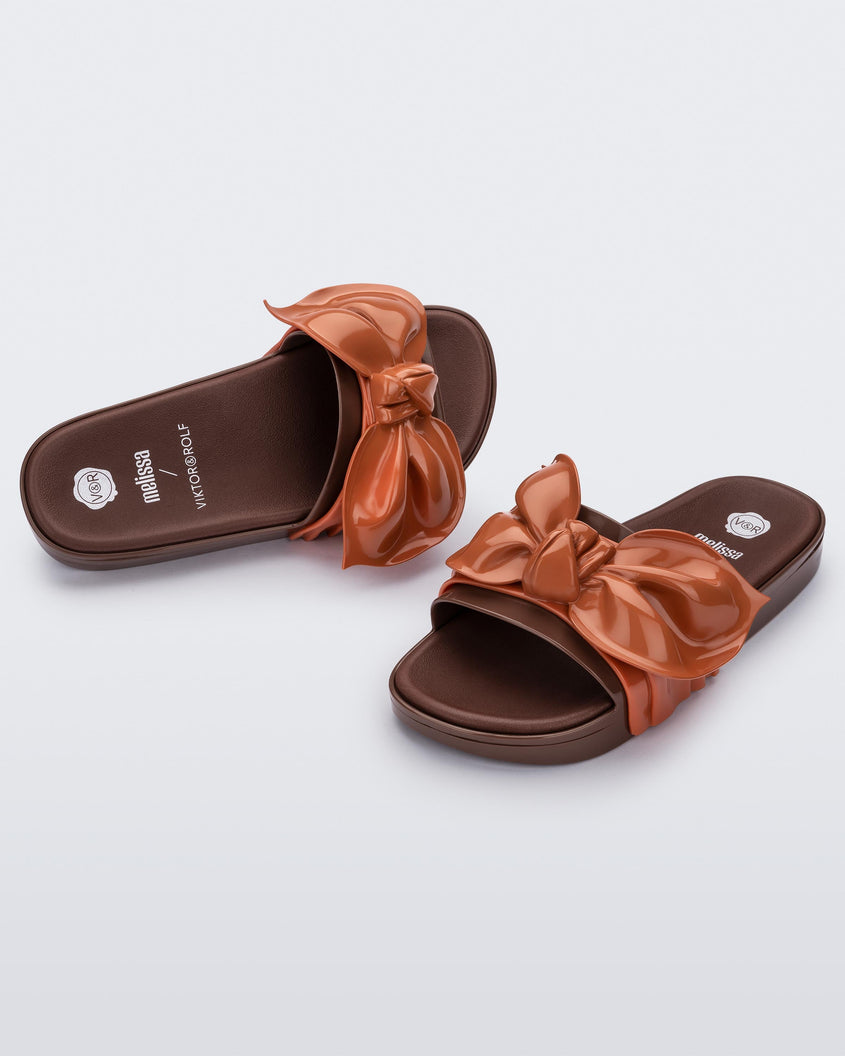 Angled view of a pair of Melissa Tie Beach slides with brown sole and brown/orange strap with 3D bow detail. 
