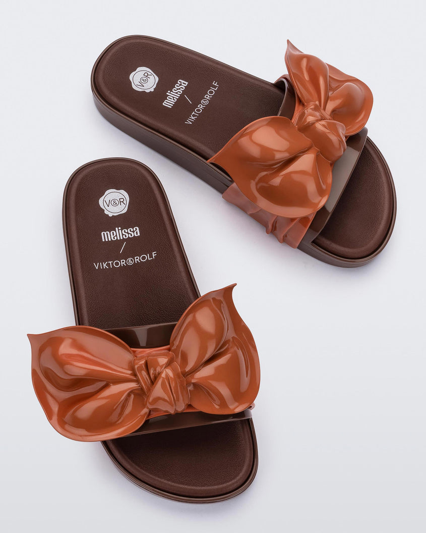 Top view of a pair of Melissa Tie Beach slides with brown sole and brown/orange strap with 3D bow detail. 