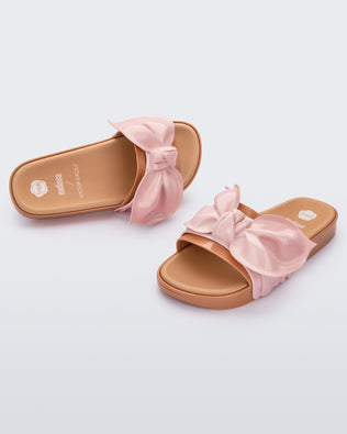 An angled front and top view of a pair of beige/pink Melissa Tie Beach slides with a beige base and a pink bow on the top strap.