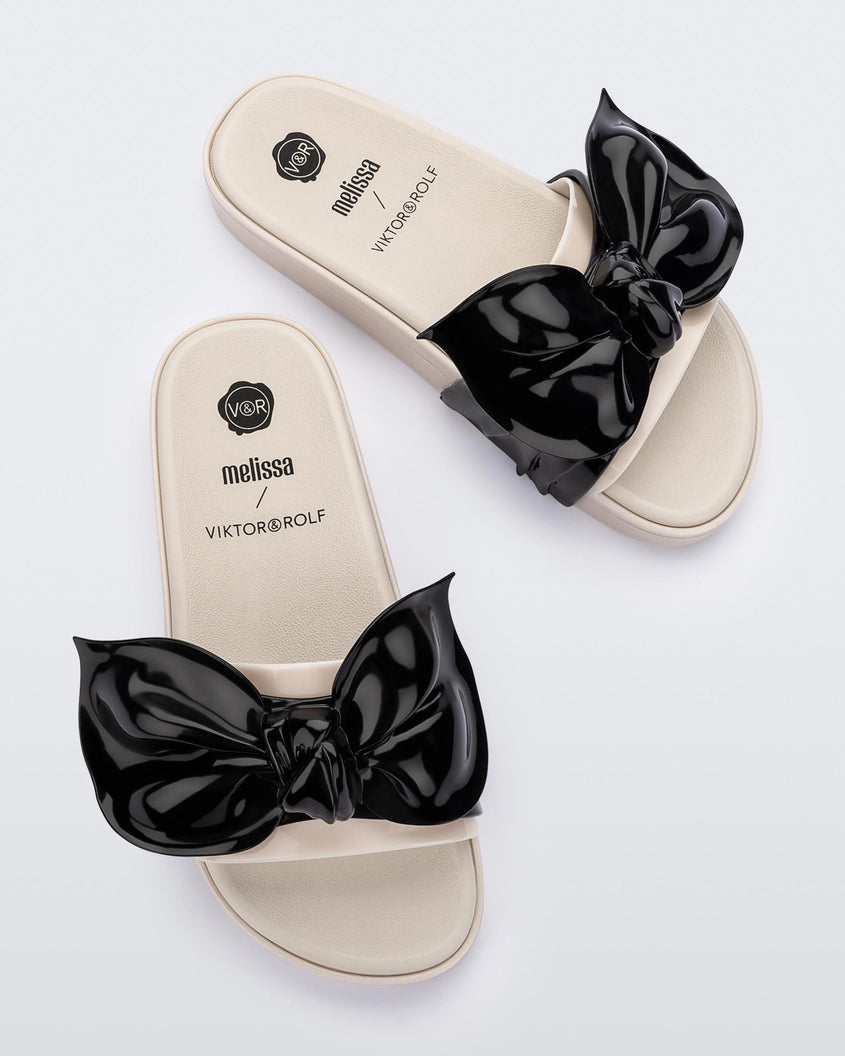 Top view of a pair of Melissa Tie Beach slides with beige sole and beige/black strap with black 3D bow attached. 