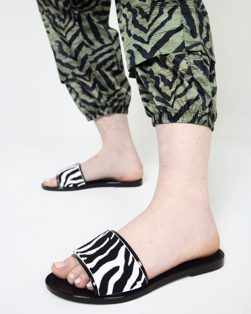 A model's legs in green pants and a pair of green/black pants and a pair of black/white Melissa Savage slides with a zebra print on the strap.