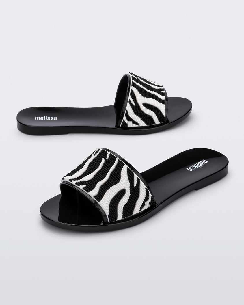 An angled side view of a pair of black/white Melissa Savage slides with a zebra print on the strap.