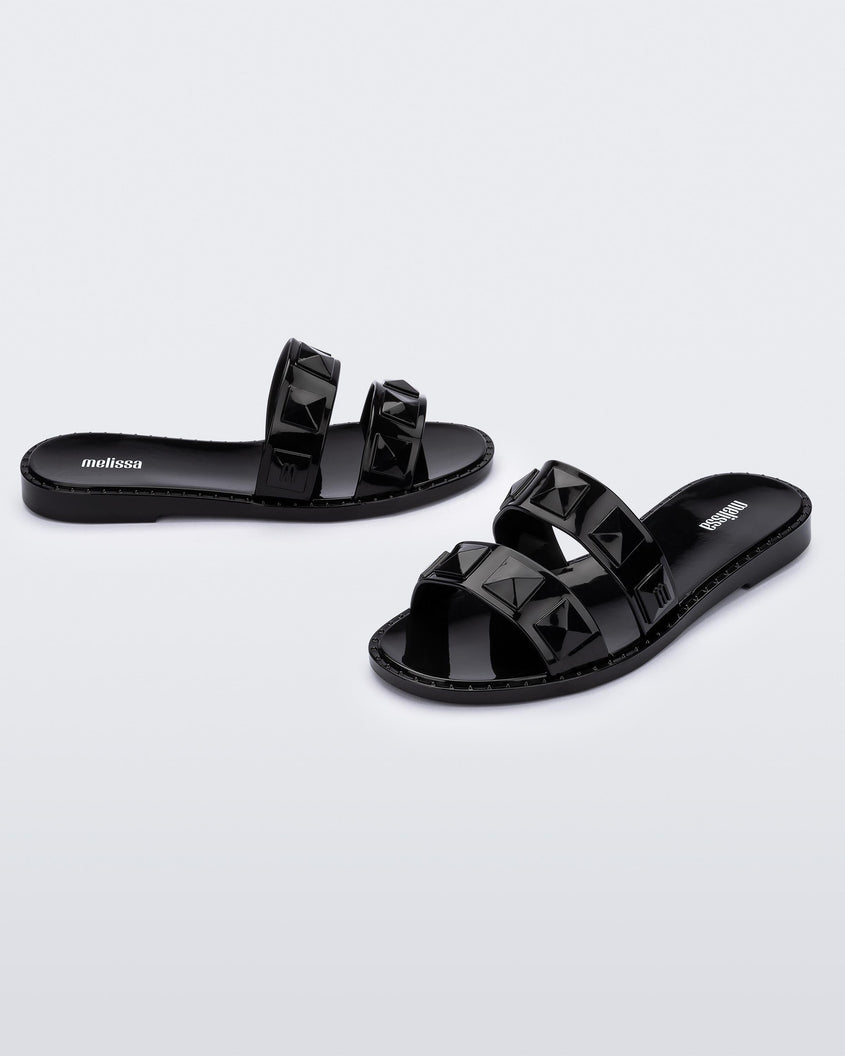 An angled front and side view of a pair of black Melissa Lucy slides with stud details on the two straps.