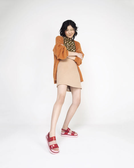 A model posing for a picture in a brown coat and a pair of transparent red/pink Melissa Cosmic platform sandals with a pink sole and two red straps.
