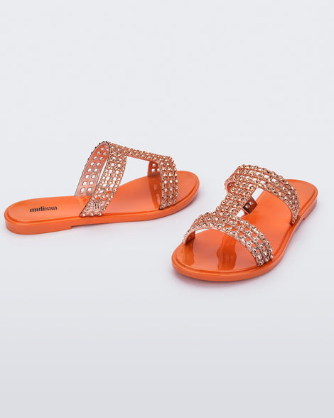 An angled front and side view of a pair of orange / copper Melissa Glowing slides with copper gem stones on the straps and an orange sole.