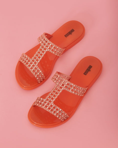 Top view of a pair of orange / copper Melissa Glowing slide with copper gem stones on the straps and an orange sole, on a pink background.