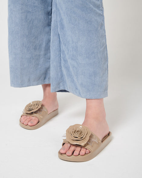 A model's legs in blue corduroy pants and a pair of beige Melissa Spikes Beach slides with a flower detail and spikes on the front strap.