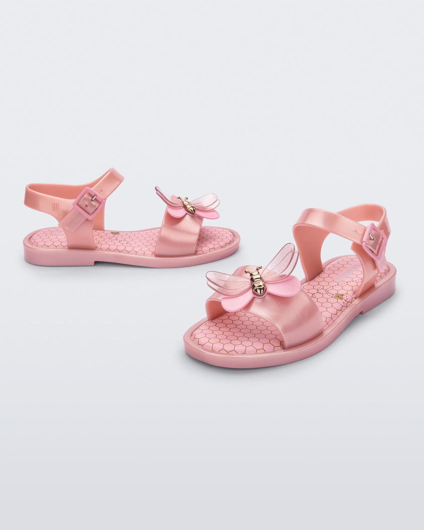 An angled front and side view of a pair of a pearly pink Mini Melissa Mar Bugs sandal with a honeycomb pattern insole and a pink bug detail with a gold buckle on the front.