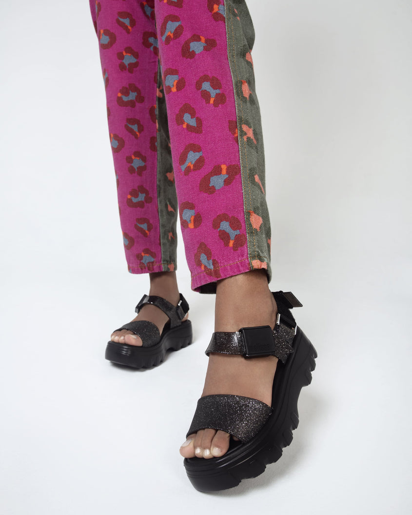 A model's legs wearing a pair of black/glitter black platform Melissa Kick Off Sandal with two glitter straps.