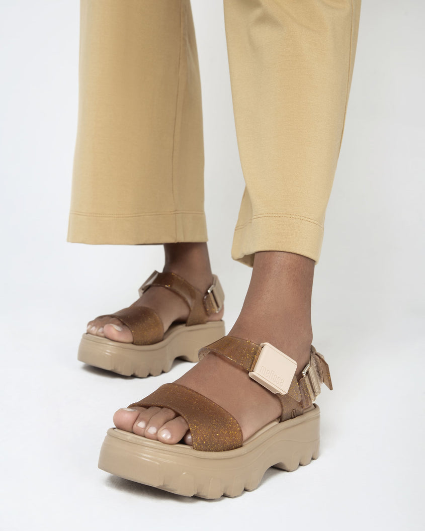 A model's legs in yellow pants wearing a pair of beige/gold platform Melissa Kick Off Sandal with two glitter straps.