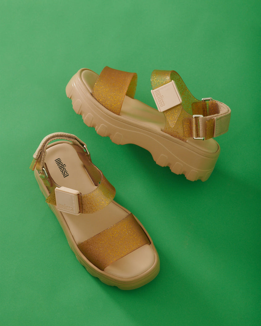 An angled top and side view of a pair of beige/gold platform Melissa Kick Off Sandals with two glitter straps on a green surface.